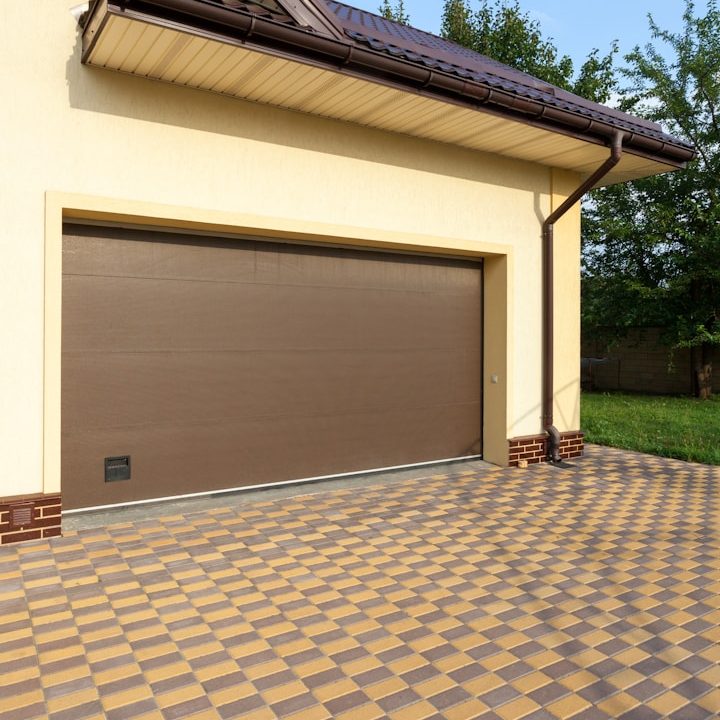 Porte de garage enroulable marron, maison avec allée en carreaux, jardin à l'arrière.